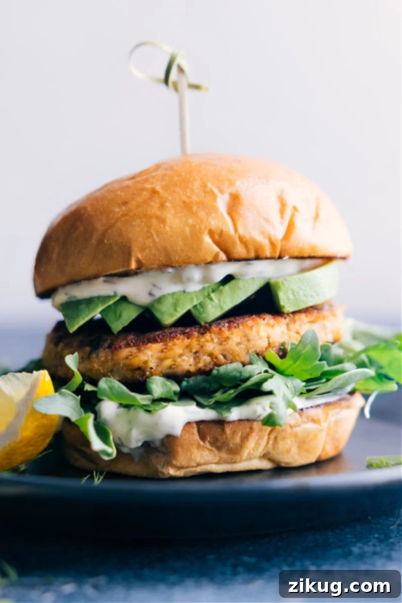 A perfectly cooked salmon burger served on a toasted brioche bun, generously topped with fresh greens, creamy avocado slices, and a dollop of zesty tartar sauce, presented on a plate.