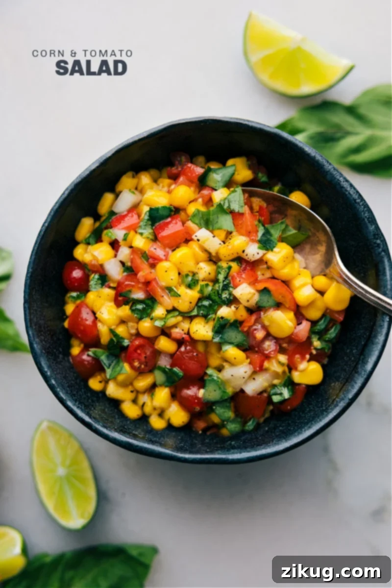 This Corn and Tomato Salad recipe features a vibrant blend of sweet, juicy corn and tomatoes balanced by the crunch of diced bell pepper and onion. The tangy lime vinaigrette, spiked with a hint of maple syrup and Dijon mustard, adds a burst of citrusy freshness that ties together everything beautifully! A perfect summer side dish via chelseasmessyapron. Overhead image of a vibrant Corn and Tomato Salad in a white bowl, showcasing golden corn kernels, halved red cherry tomatoes, diced red bell pepper, and fresh green basil leaves. The salad appears fresh and appealing.
