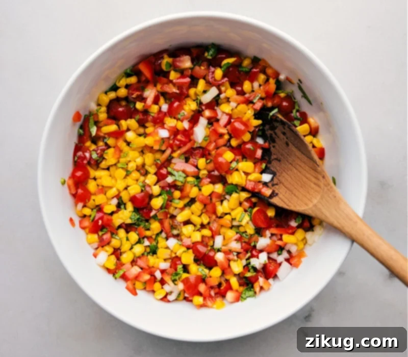 This Corn and Tomato Salad recipe features a vibrant blend of sweet, juicy corn and tomatoes balanced by the crunch of diced bell pepper and onion. The tangy lime vinaigrette, spiked with a hint of maple syrup and Dijon mustard, adds a burst of citrusy freshness that ties together everything beautifully! Recipe via ChelseasMessyApron.com #cornsalad #corntomatoes #corn #tomatoes #salad #lime #vinaigrette #summery #basil Overhead view of a vibrant bowl of Corn and Tomato Salad, garnished with fresh basil leaves, with a rustic wooden serving spoon resting in the bowl. The salad looks inviting and fresh.