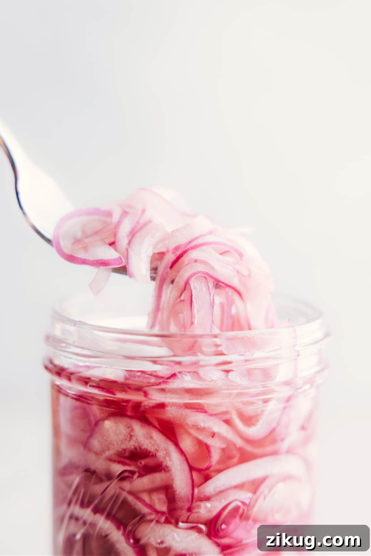 The Best Easy Pickled Red Onions A fork lifting vibrant pink pickled red onions out of a glass jar, ready to be served.