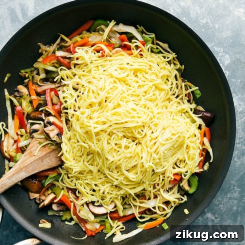 Freshly cooked noodles being added to a large skillet filled with vibrant, stir-fried vegetables like cabbage, carrots, and bell peppers, ready to be combined for a delicious Chow Mein.