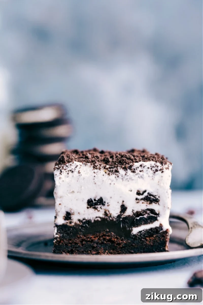 This Brownie Oreo Ice Cream Cake is an irresistible frozen dessert featuring fudgy brownies, hot fudge, chopped Oreos, and cookies & cream ice cream. A delectable slice of Brownie Oreo Ice Cream Cake on a plate, showcasing its distinct layers of brownie, Oreos, hot fudge, ice cream, and whipped cream.