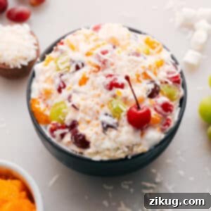 Close-up of a vibrant Ambrosia Salad in a decorative glass bowl, showcasing the creamy dressing, colorful fruits like pineapple and mandarin oranges, and fluffy mini marshmallows, garnished with a cherry.