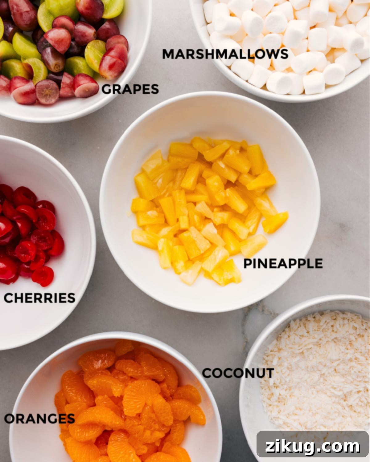 Ingredients Prepped Out For The Best Ambrosia Salad All the vibrant ingredients for Ambrosia Salad laid out on a white surface: canned pineapple tidbits, mandarin orange segments, halved green grapes, red maraschino cherries, white and rainbow mini marshmallows, and shredded coconut flakes, meticulously prepared.