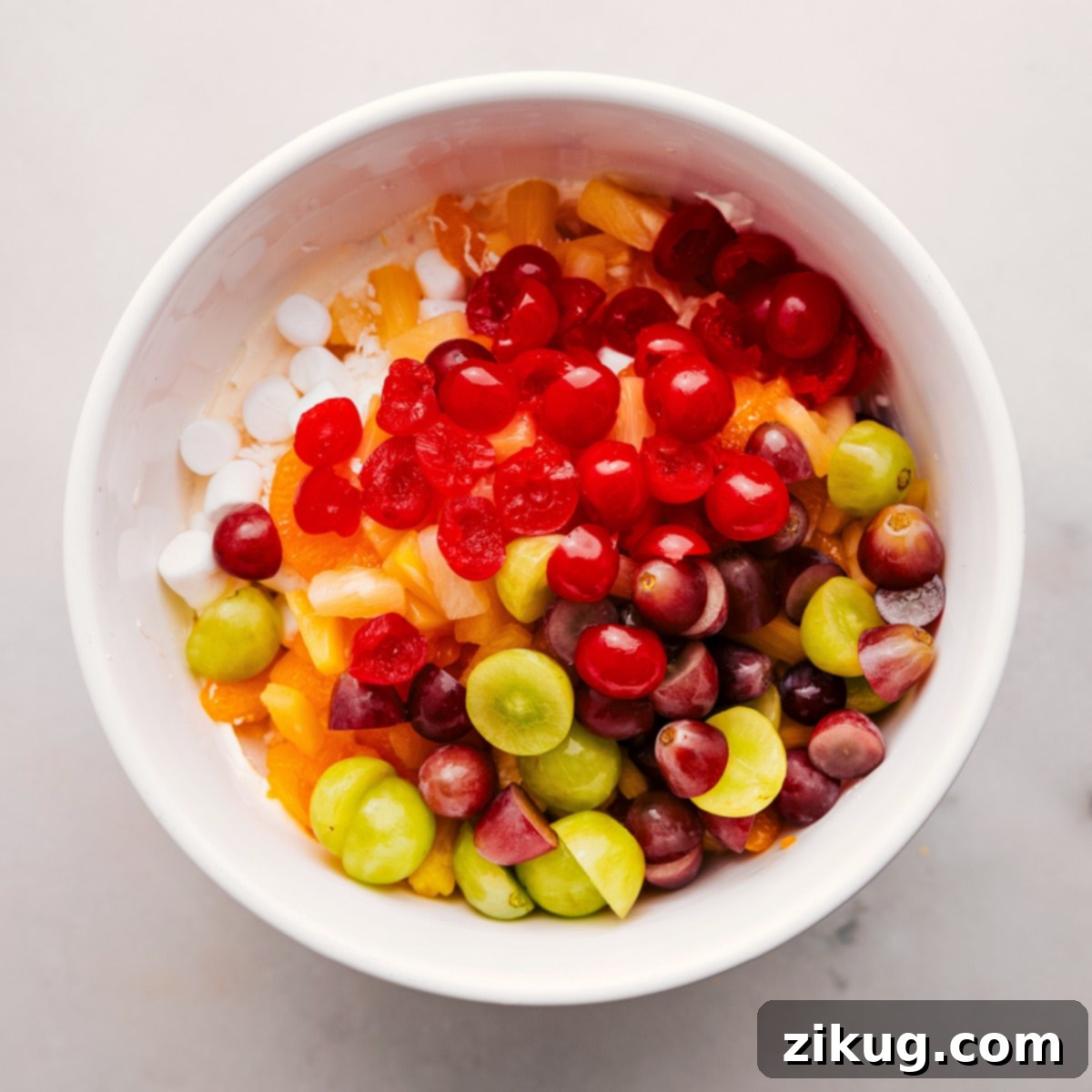 How To Make Ambrosia Salad Recipe: Fruit Base A large glass bowl filled with the colorful fruit and marshmallow base for Ambrosia Salad, showcasing vibrant mandarin oranges, crisp green grapes, bright red cherries, and fluffy white marshmallows, ready for the dressing.