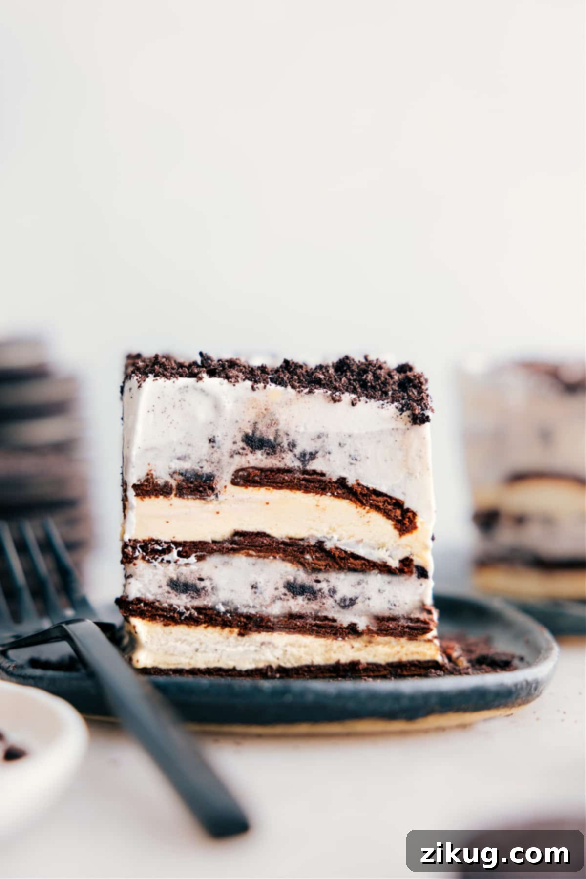 Delicious Four-Ingredient Ice Cream Sandwich Cake Slice A perfectly sliced piece of four-ingredient Ice Cream Sandwich Cake on a white plate, revealing its delicious layers of ice cream sandwiches, cookies and cream ice cream, and whipped cream with crushed Oreos.