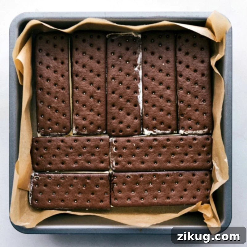 Base Layer of Ice Cream Sandwich Cake Assembly in Pan The first layer of vanilla ice cream sandwiches neatly arranged at the bottom of a parchment-lined 9x9 inch baking pan, forming the base of the frozen cake.