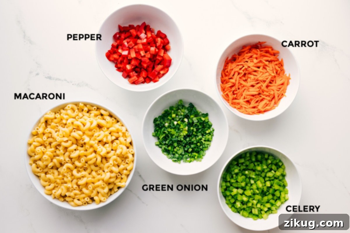 All the fresh ingredients for this Macaroni Salad Recipe, including dry elbow macaroni, vibrant red bell pepper, crisp carrots, aromatic green onions, and crunchy celery, prepped and ready for assembly on a rustic wooden board.