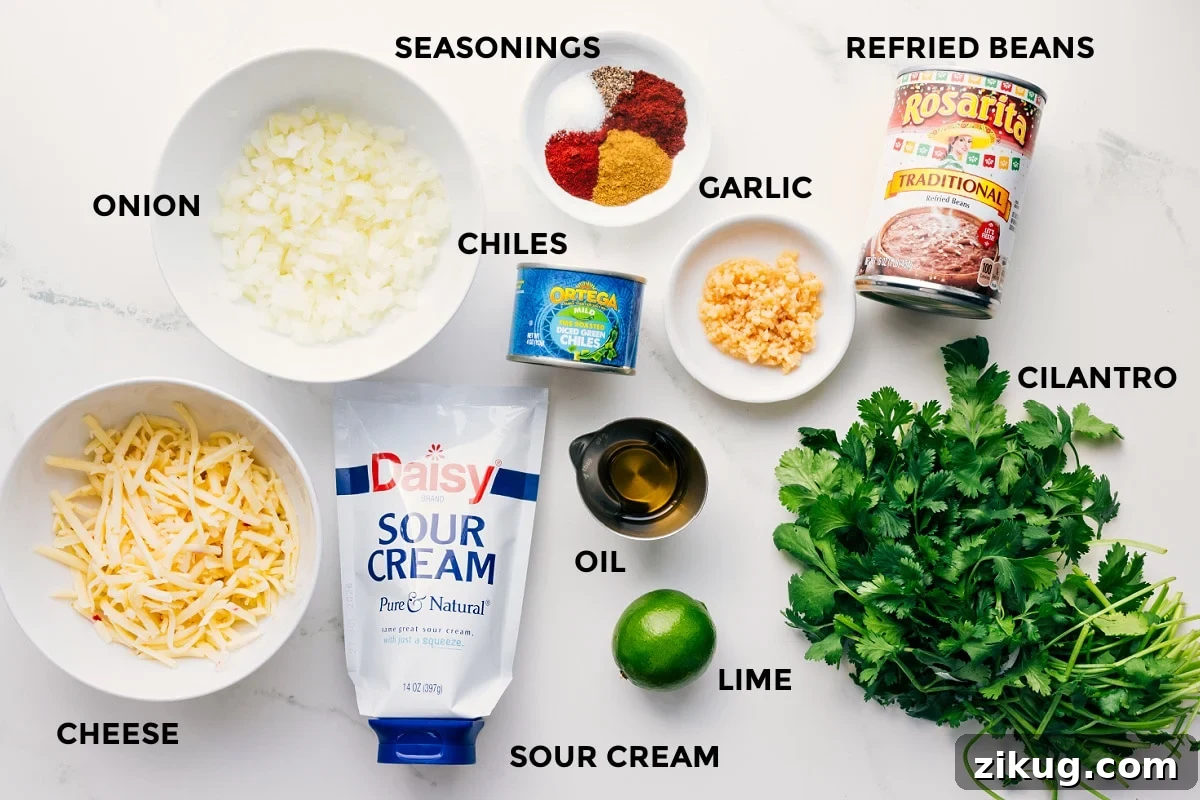 Ingredients In This Refried Bean Dip All the ingredients in this recipe prepped out for easy assembly including the cheese, onion, sour cream, lime, oil, chiles, seasoning, garlic, refried beans, and cilantro.